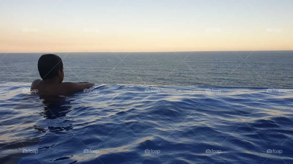 Kid looking to the vast infinity of the ocean at borderless pool over sunset.