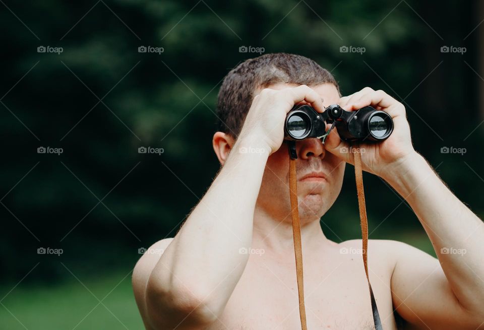 Portrait of a young handsome Caucasian male brunet who looks very attentively and seriously through binoculars, standing in a park on a summer day, close-up view from the side.