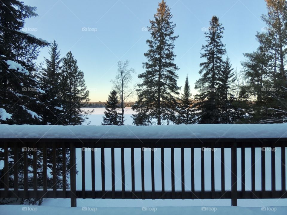 Snow covered porch railing overlooking pine trees and a lake 