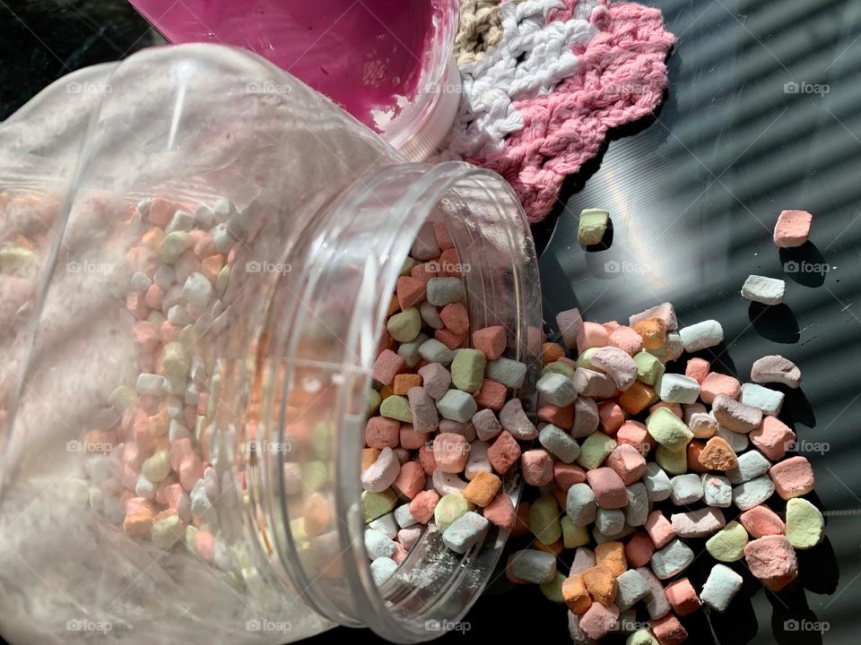 Colorful Pastel Mini Marshmallows Falling Off Clear Plastic Jar By Pink Slime Container On A Crocheted Cloth.