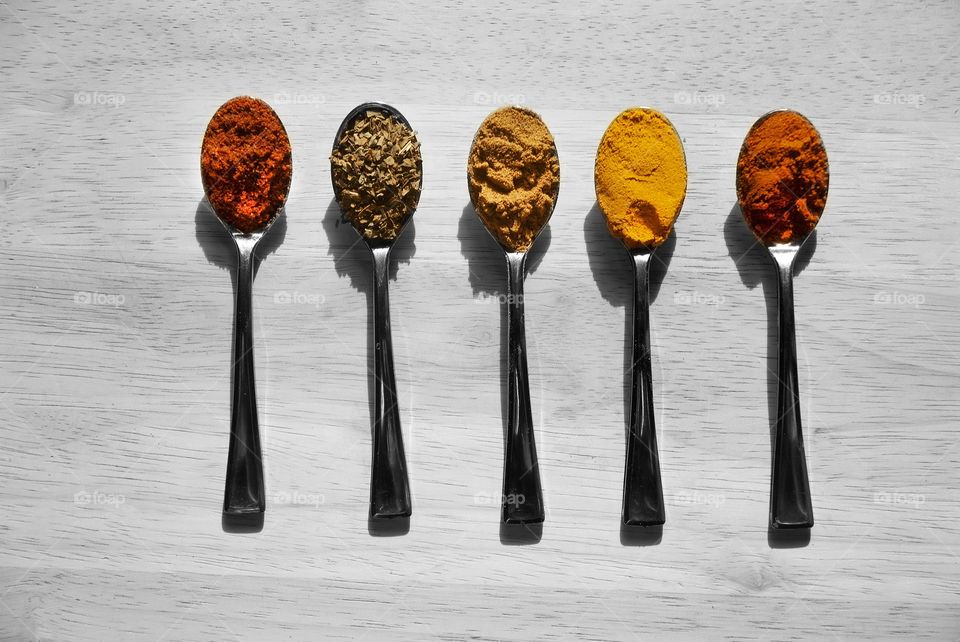 Mini spoons with tandoori masala, curry, basil, turmeric, and cayenne powder respectively in each are arranged on a black and white canvas wooden cutting board.