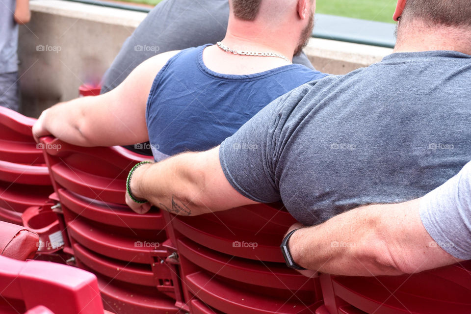 From behind view of three men sitting in stadium seats outdoors with each of their arms around the back of the next person's chair