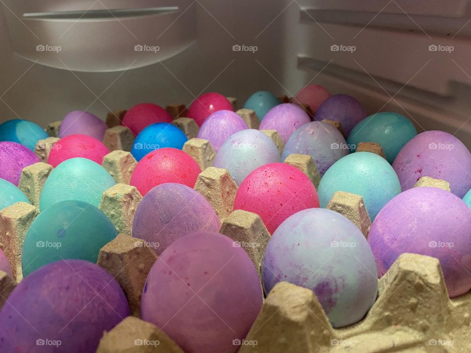 A variety of pastel, dye-colored Easter Eggs drying on an egg carton on the refrigerator shelf. The eggs are pink, purple, green, turquoise, blue, and teal. 