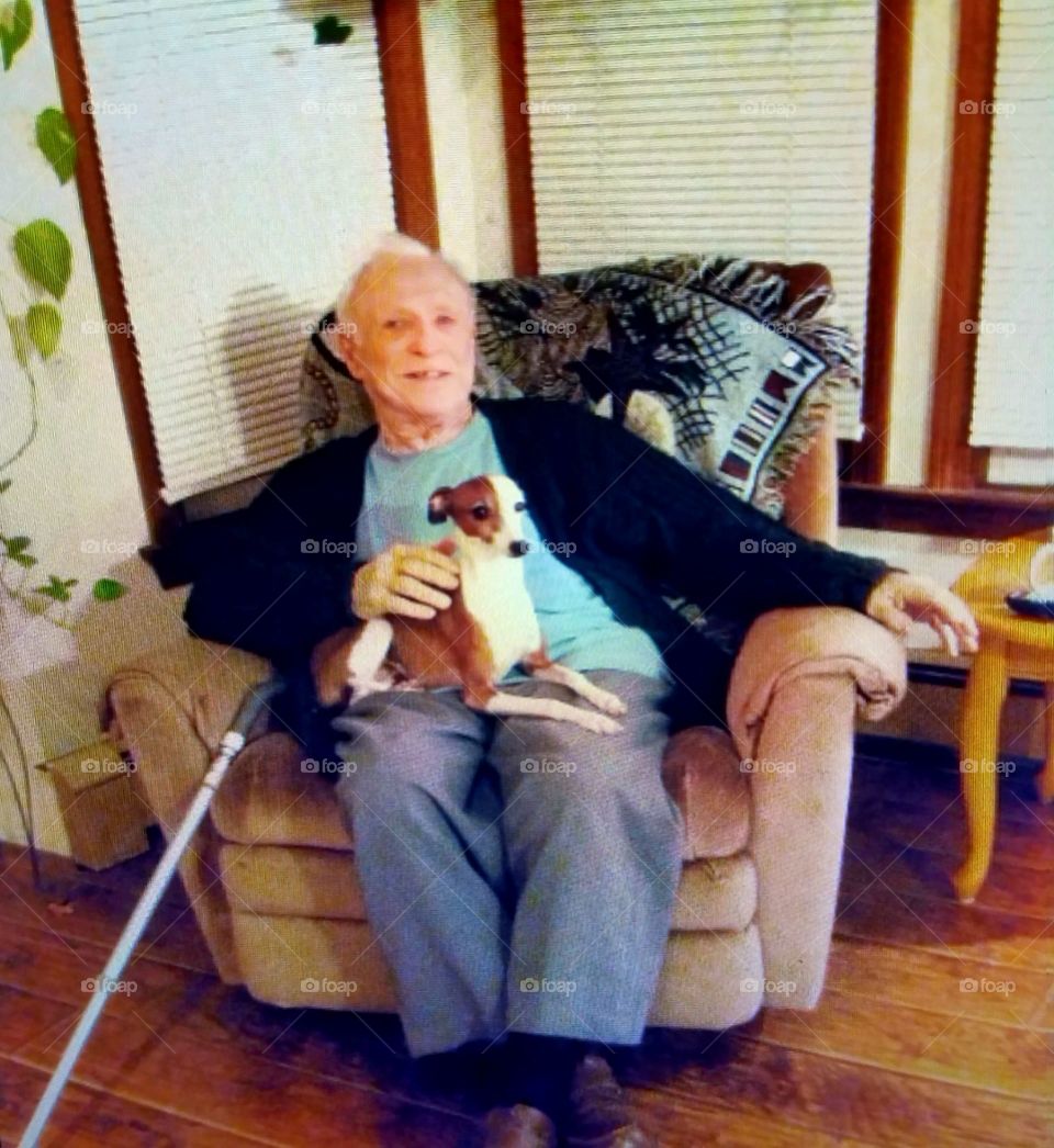 Older man portrait in chair with cane & dog posed on his lap.