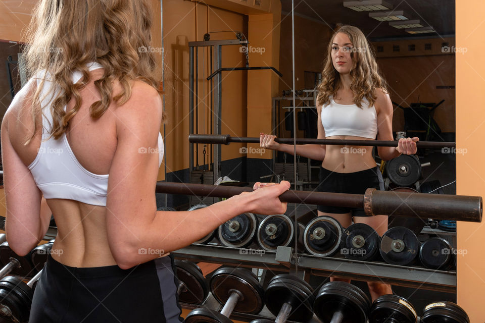 Young and beautiful woman looking in the mirror and working out with dumbbells in gym.