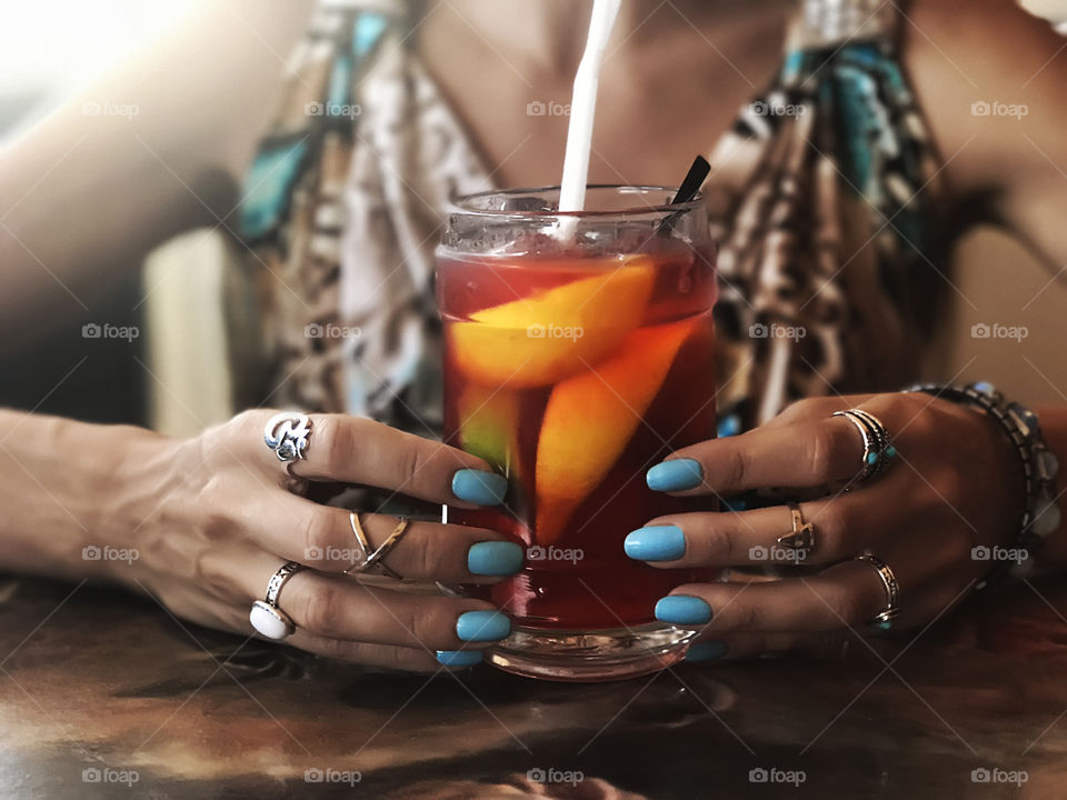 Young woman in colorful summer dress with blue nail polish and fashionable accessories drinking citrus colorful summer cocktail 