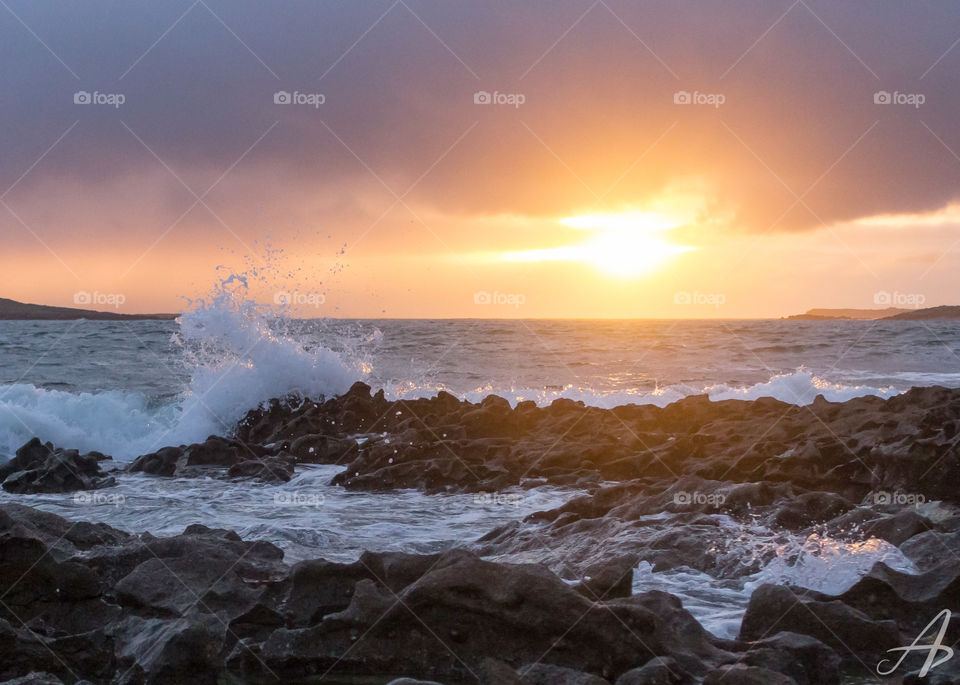 A wave crashes on he rocks as the golden sun gets ready to set