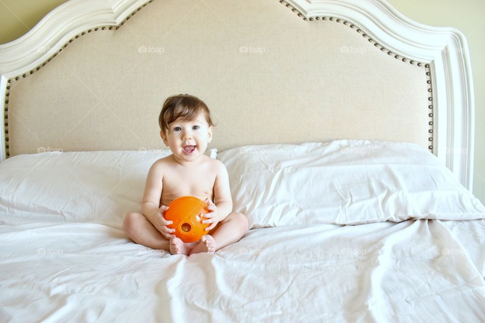 Smiling young toddler holding an orange ball and sitting on a bed with fresh clean white sheets 