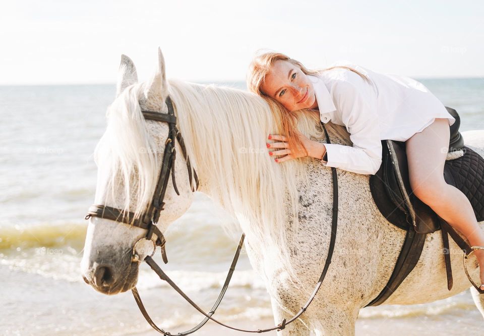 Young long hair woman in white shirt with white horse on seascape background