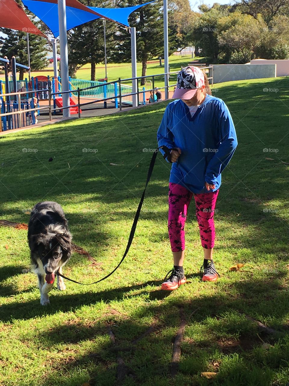 A young girl wearing pink leggings and active wear walking her border collie sheepdog on a leash in a park near a playground