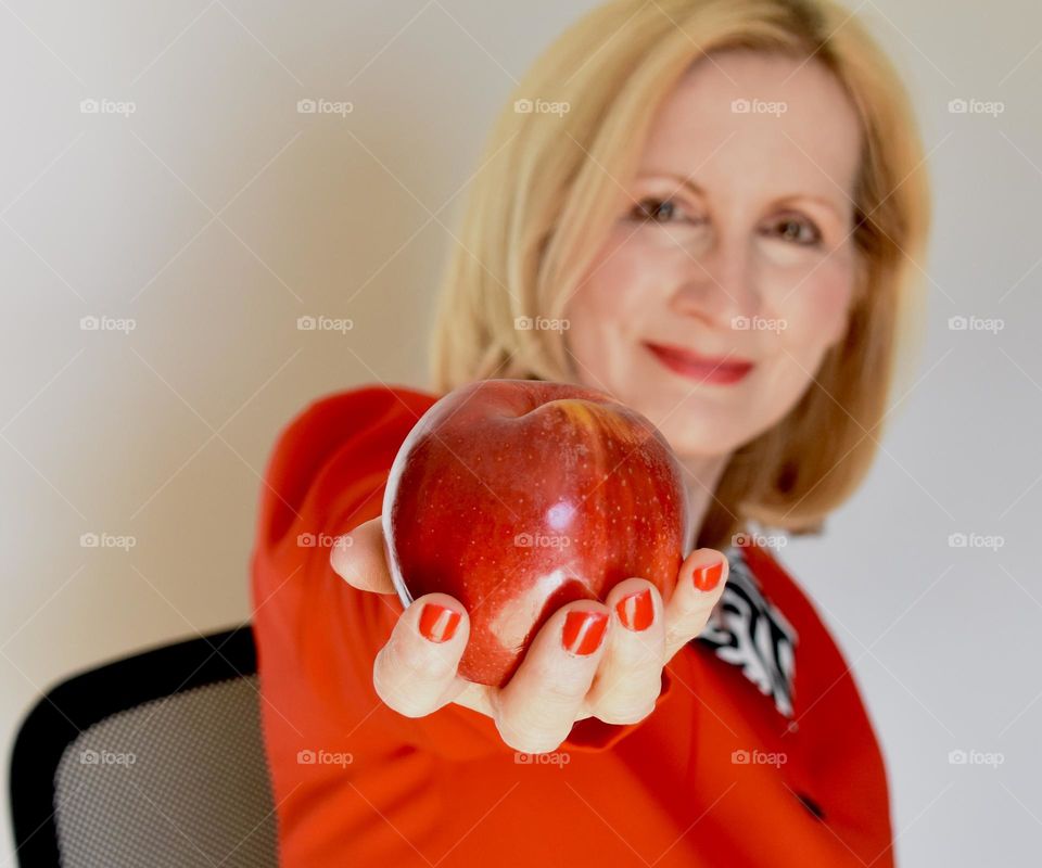 Blonde woman in a red suit holding out a red apple