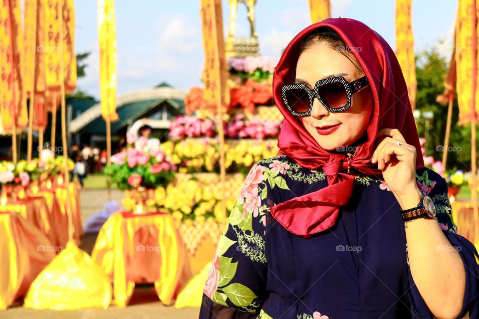 Portrait of a beautiful mother wearing a red hijab, wearing a floral patterned robe and wearing black glasses, taking a photo with a happy expression.