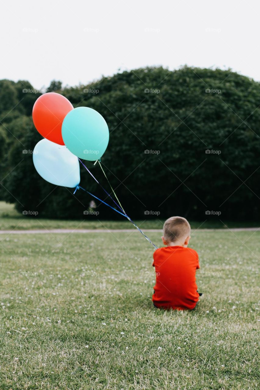 A little blond boy in a red T-shirt sits on a green field, holding a bunch of colorful balloons in his hand, back view 