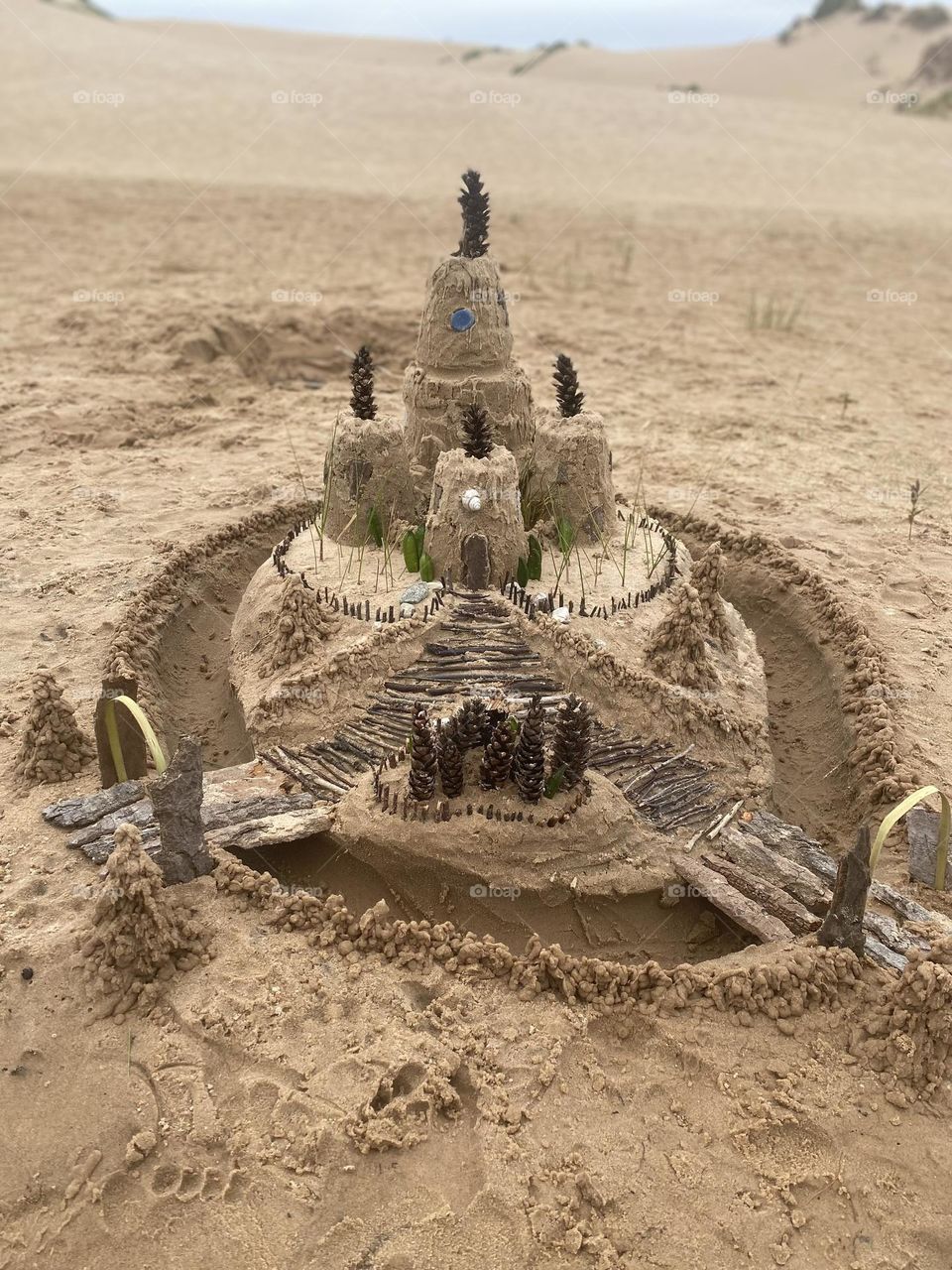 This image shows an intricate sandcastle on a beach, featuring multiple tiers and decorated with natural elements like pinecones, sticks, leaves, and shells. The sandcastle has a central tower surrounded by smaller structures, with detailed pathways 