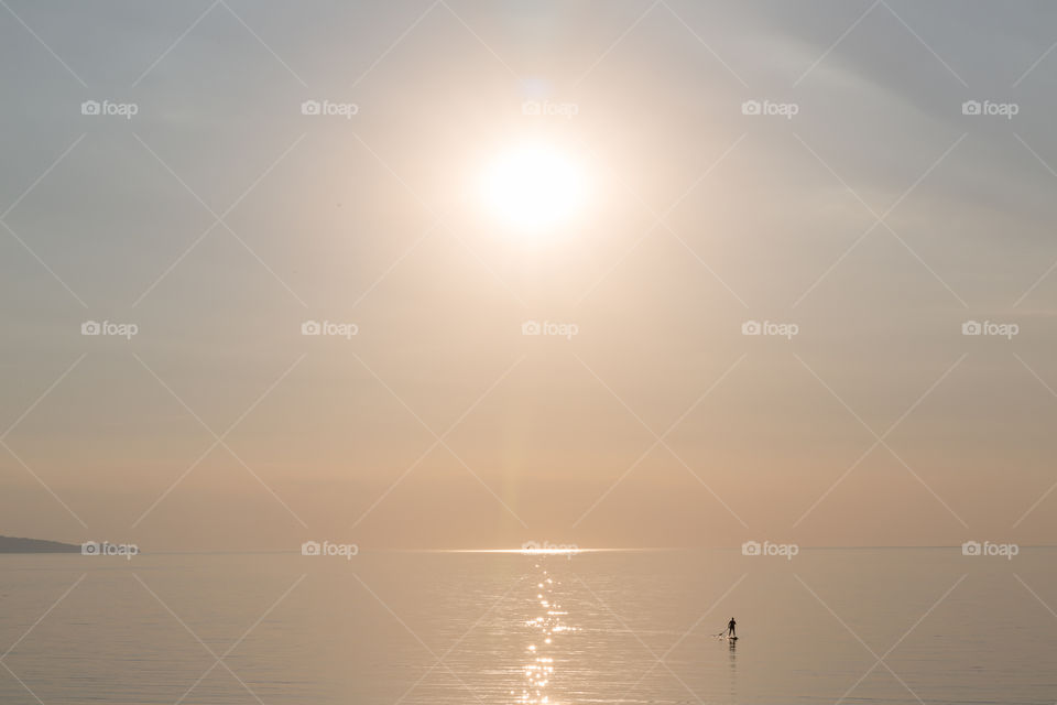 Silhouette of a man stand up paddle boarding in bright sunlight on calm sea 