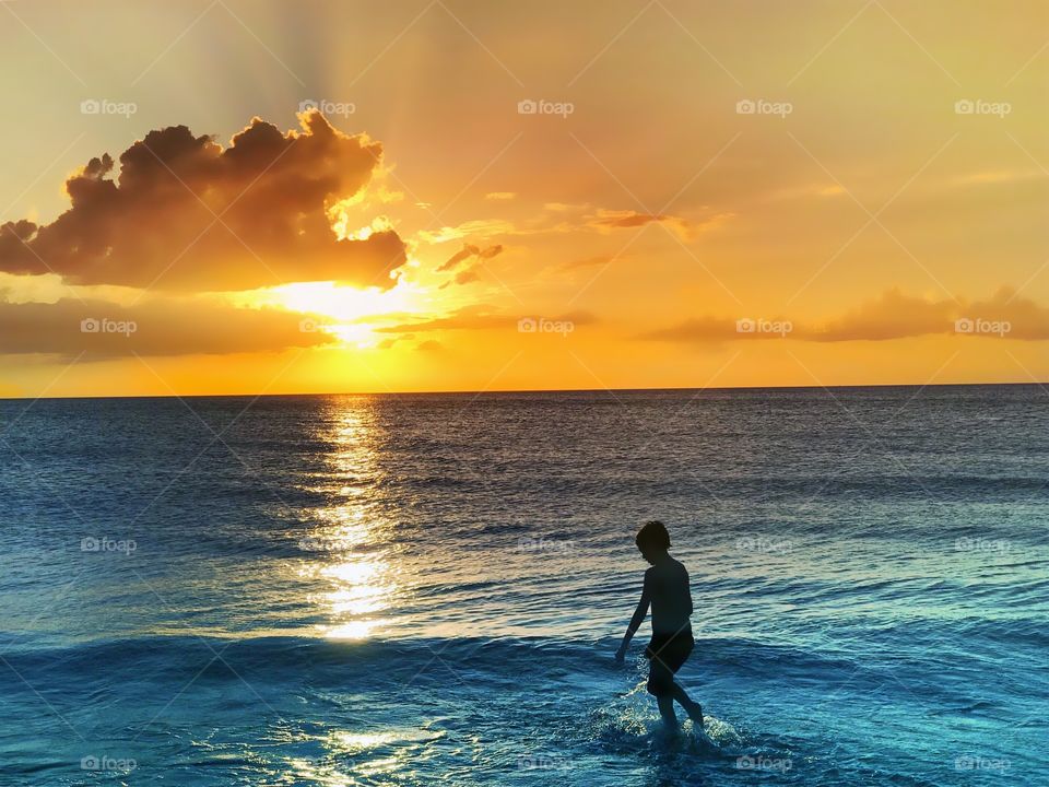 Small boy playing in the indigo ocean waves surrounded by a glorious golden sunset.