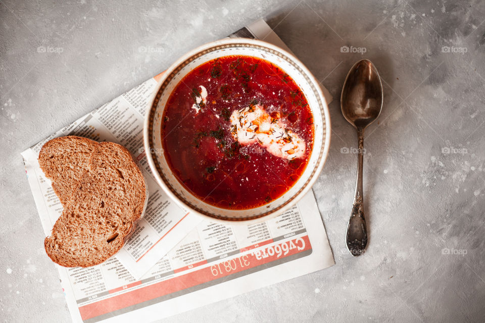 Plate of Ukrainian borsch. Nearby are two sliced pieces of bread and a spoon. A plate is on the newspaper. Photo taken on a white concrete background.Red borsch, add sour cream and greens.