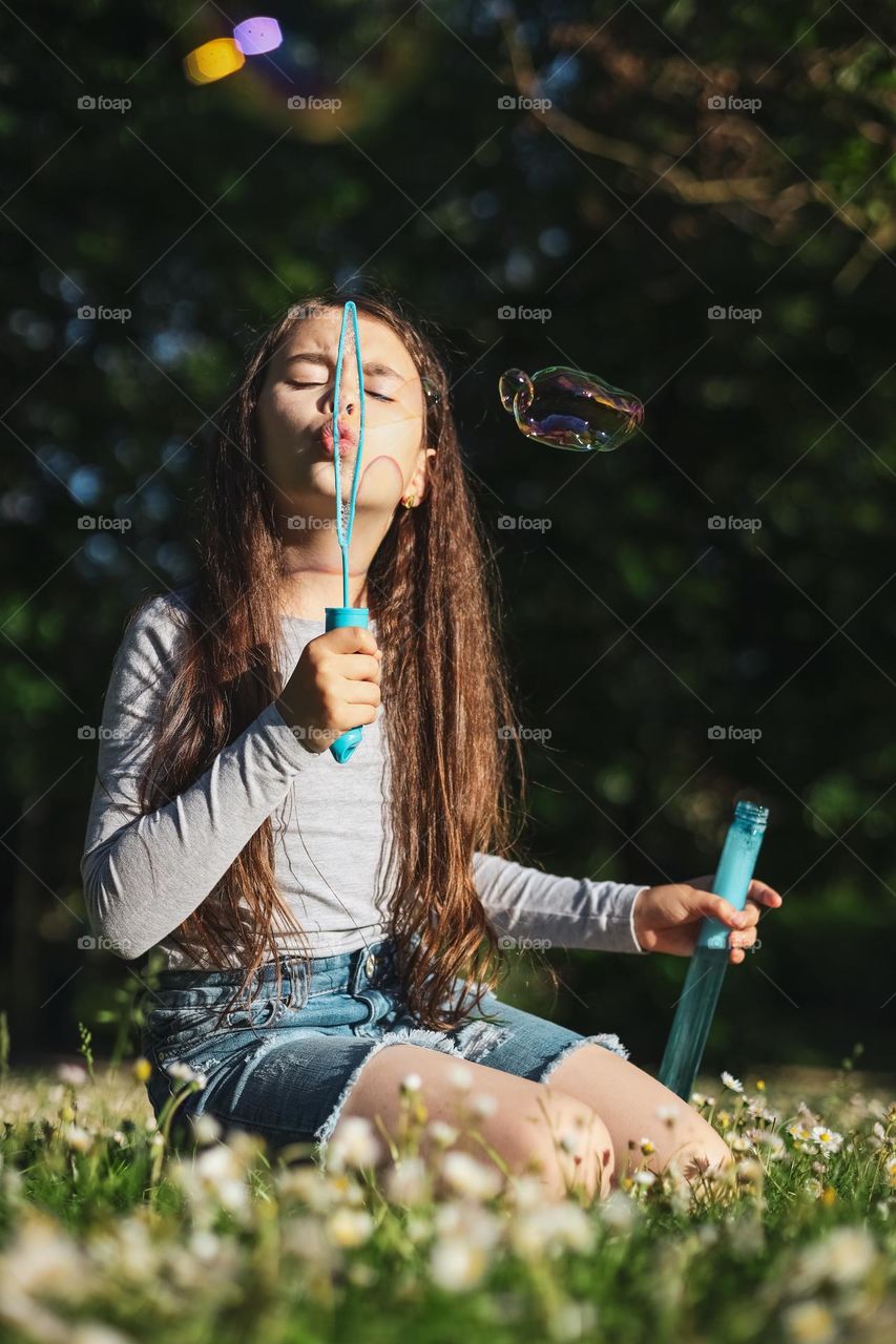 Portrait of a beautiful caucasian brunette girl with flowing hair sitting on the lawn in the park and blowing soap bubbles with closed eyes and a happy emotion on her face, close-up side view. Happiness moment concept.