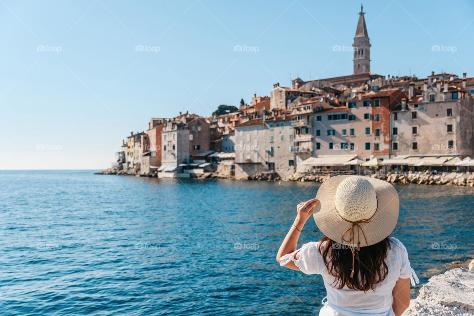 Rear view of woman wearing white dress and sun hat standing on shore of sea, admiring beautiful city of Rovinj in Croatia