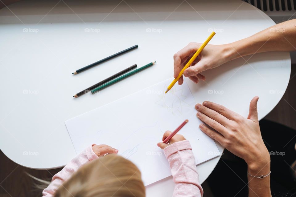 Little girl toddler with her mother drawing with colored pencils on table in children's room at home, view from top
