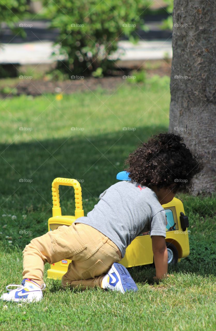 Toddler boy with Afro playing with yellow Little Tikes car in park in June 