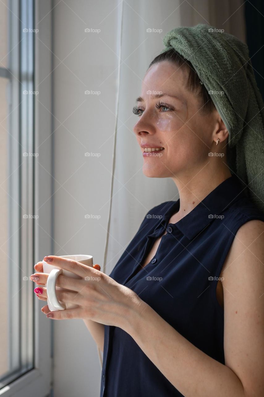 young girl at home after a shower looks out the window holding a mug of her hands with a beautiful manicure