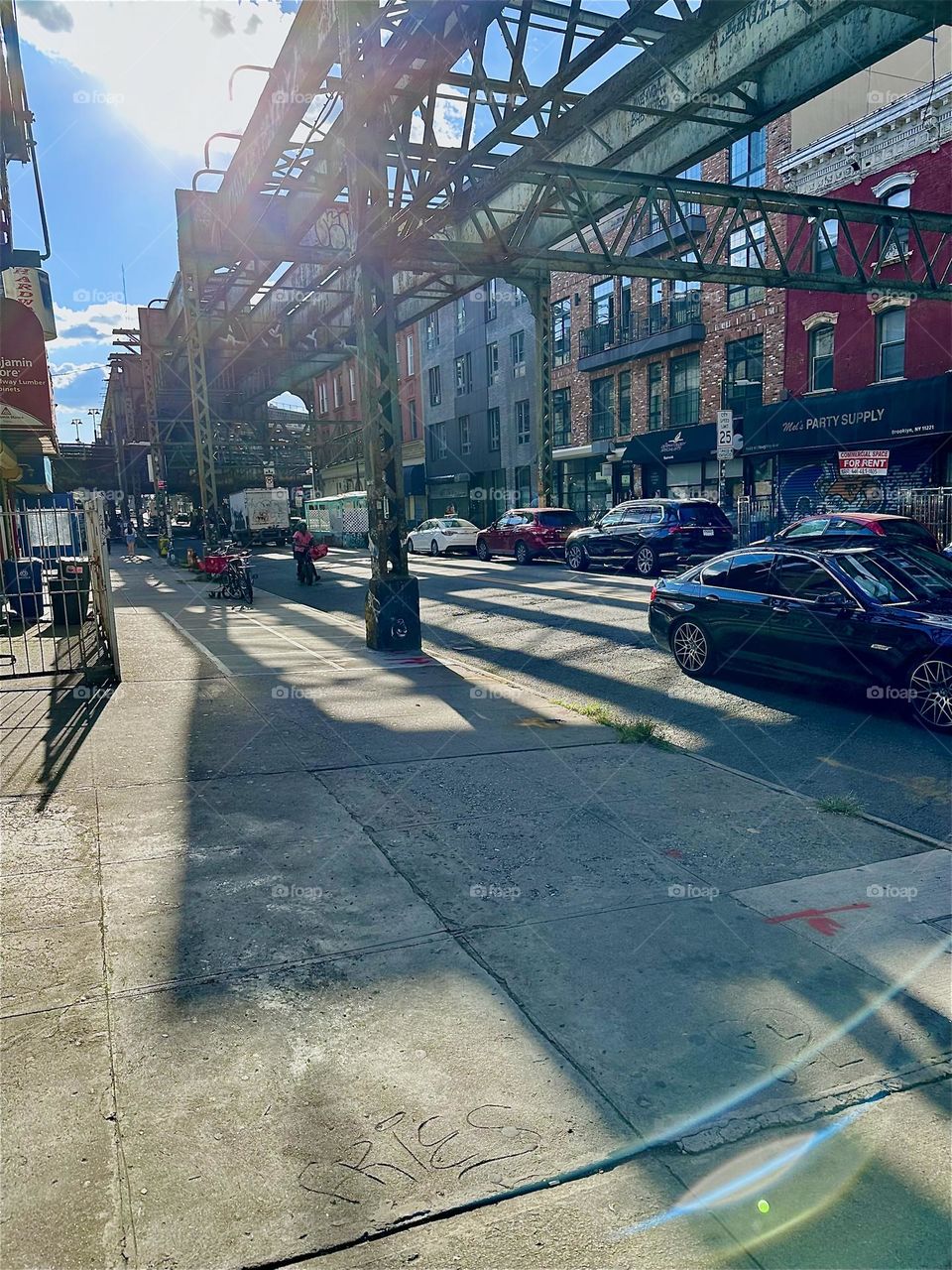 This is “Myrtle Ave” in “Bushwick”, Bklyn with an elaborate early industrial age overpass extending into the distance. The incoming afternoon sun casts elongated shadows from its multifaceted structures onto the pavement. 2024. Hypnotic Productions