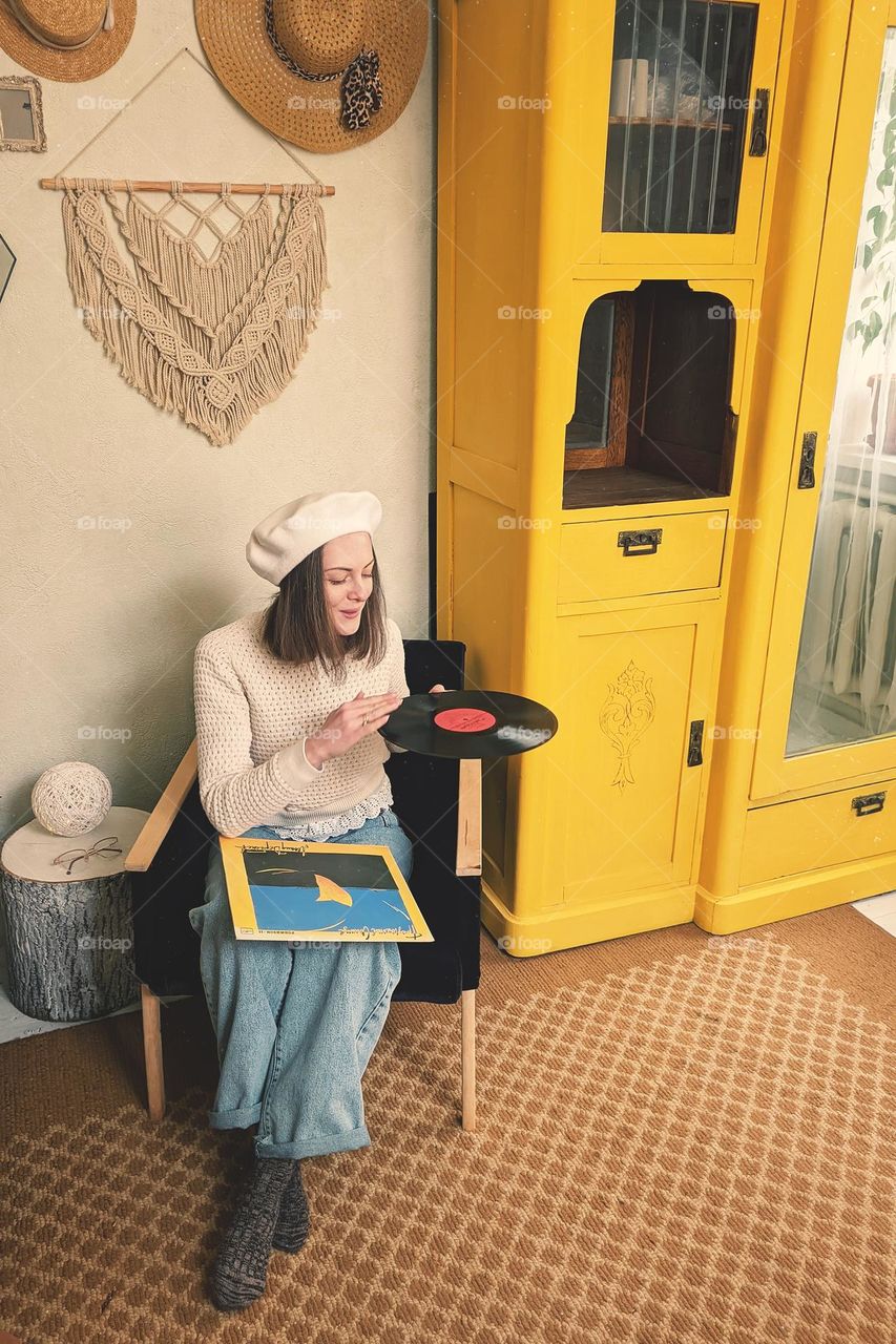 Young woman in beret sitting at home in room with music record in hands. Vintage Yellow cabinet.  Home interior