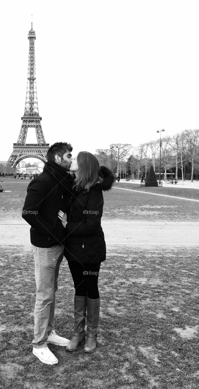 A black and white image of a couple kissing in front of the Eiffel Tower in Paris. A couple in love. Lovers kissing. Romance and togetherness. 