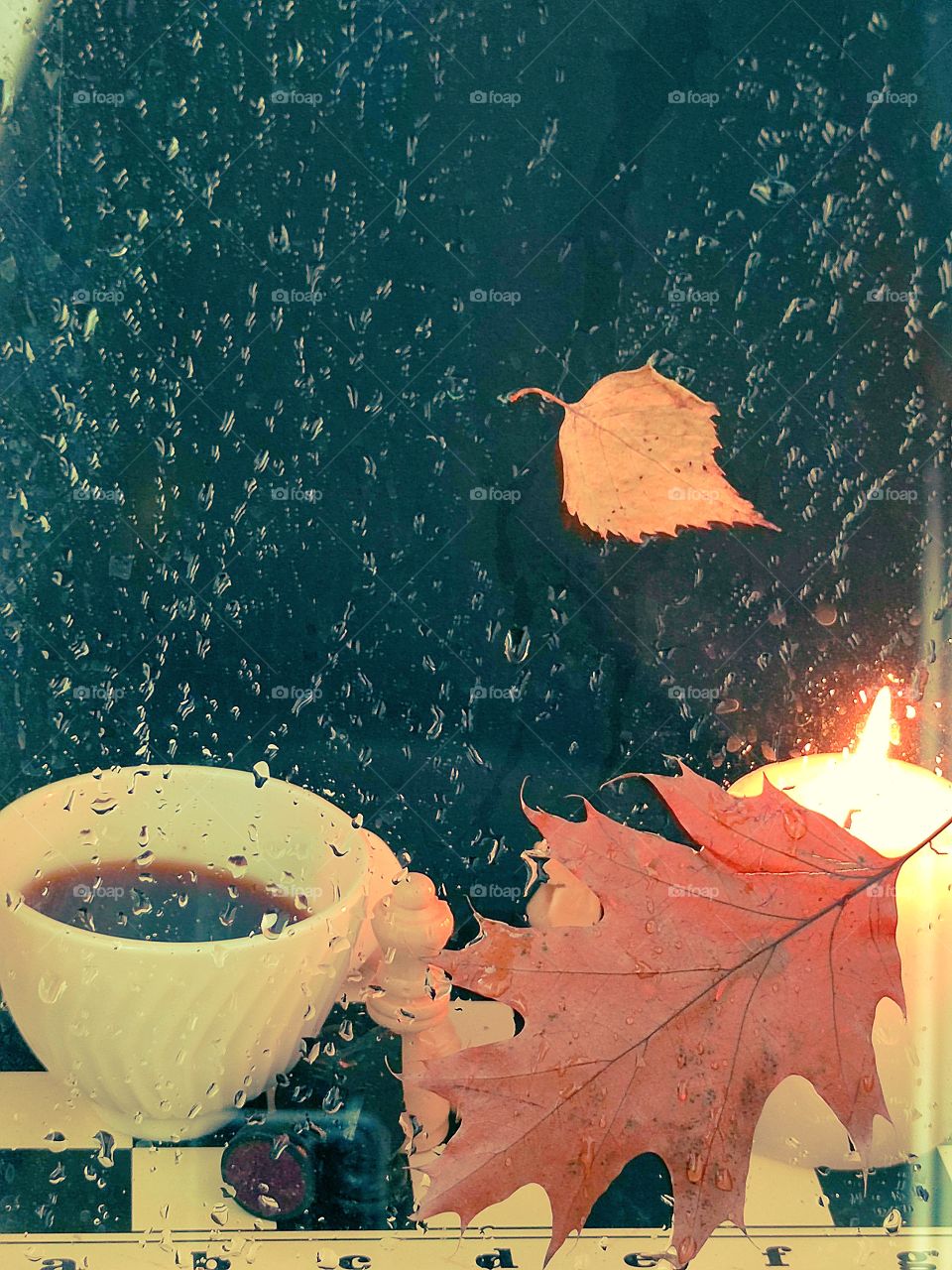 Rain. Evening. Glass in drops of water. Behind the glass is a white cup with black coffee, which stands on a chessboard, a chess piece and a burning candle. Autumn leaves stuck to the glass