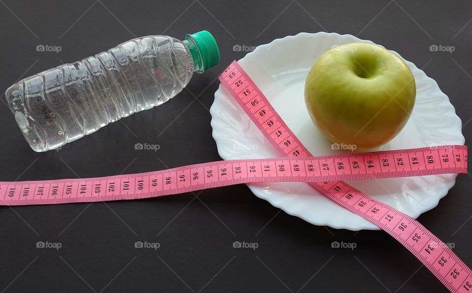 Diet concept. A green apple on a white plate tied with a measuring meter and a bottle of water on a dark background.