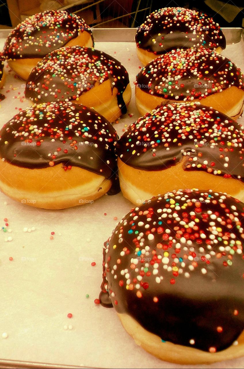 Many big round ball-shaped donuts/doughnuts with chocolate glazure and 
colorful small candies on in bakery for sale