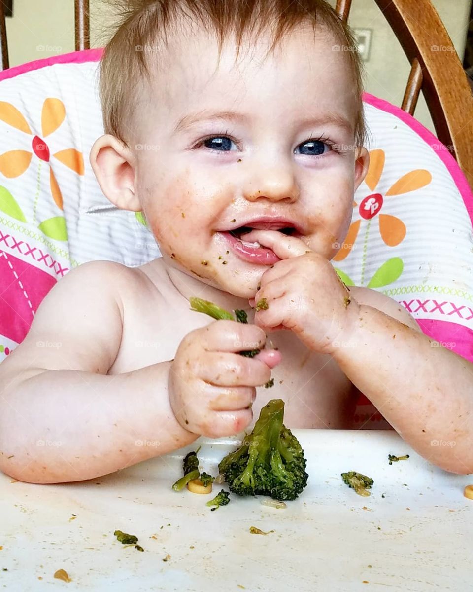 baby girl grinning and eating broccoli in high chair