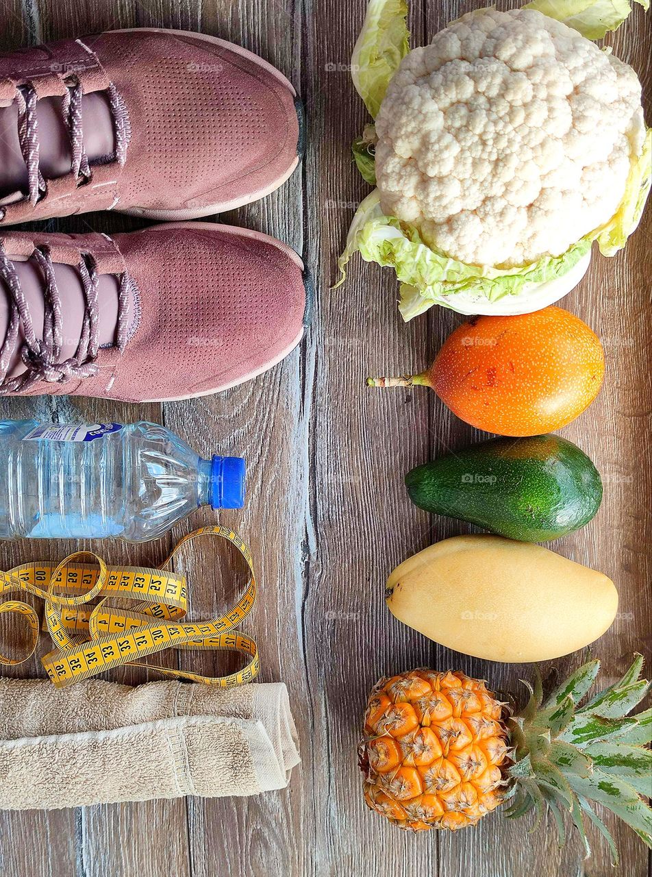 The wooden background is invisibly divided: on the one hand, fruits and vegetables, on the other hand, healthy lifestyle accessories.  Cauliflower, granadilla, avocado, mango and pineapple.  Sneakers, measuring tape, towel, bottle of water