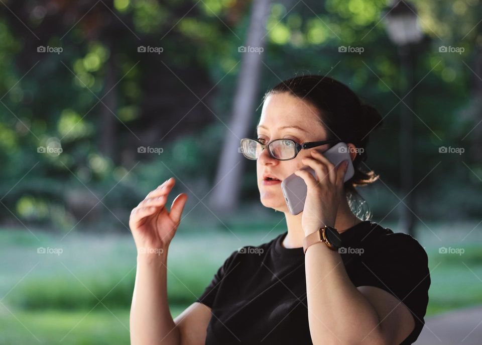 Portrait of a beautiful young caucasian brunette girl in glasses and a black t-shirt communicates very emotionally on the phone smearing her hand and looking to the side, walking in the forest on a clear sunny summer day, side view close-up .Concept