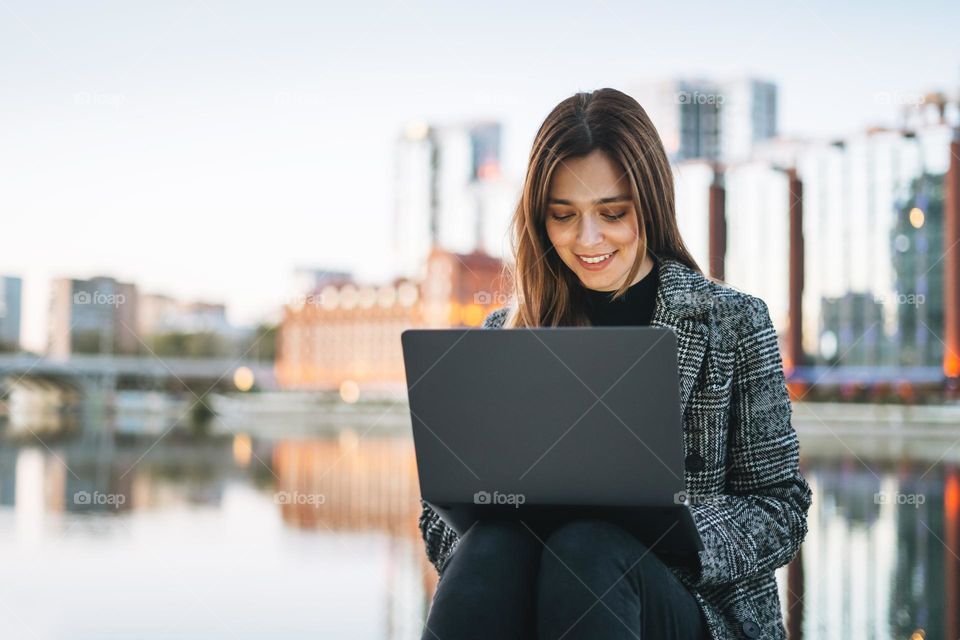 Young smiling woman in coat using laptop against evening urban city skyline