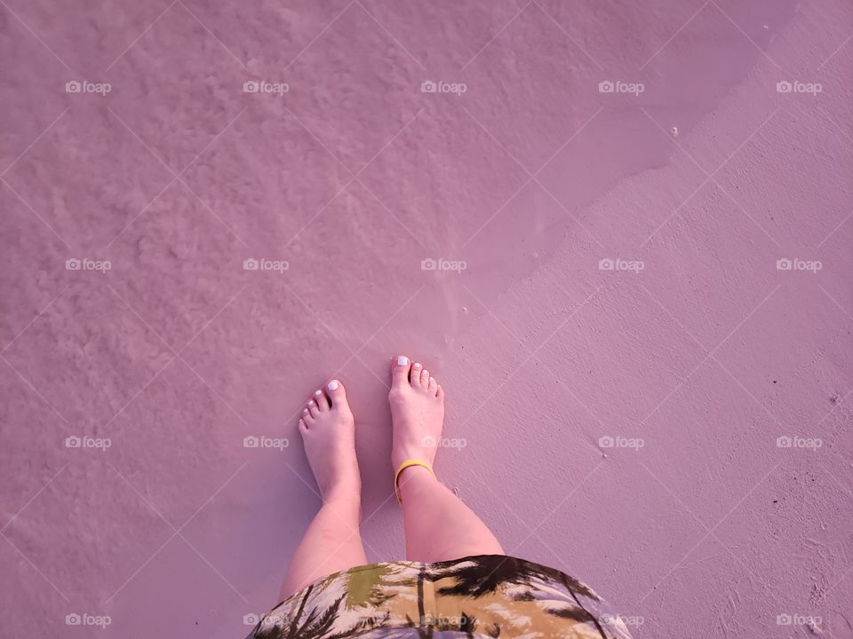 on sunny warm tropical vacation, standing on sandy beach, view looking down to feet and toes walking on pink sand and ocean water wave coming in