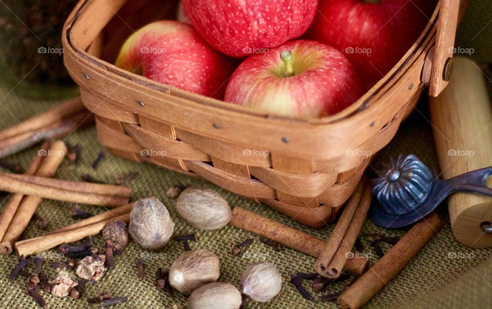 Cinnamon sticks, whole nutmeg, whole cloves, a small glass jar, a vintage pie crust crimper, a wooden pastry roller and apples in a basket on burlap