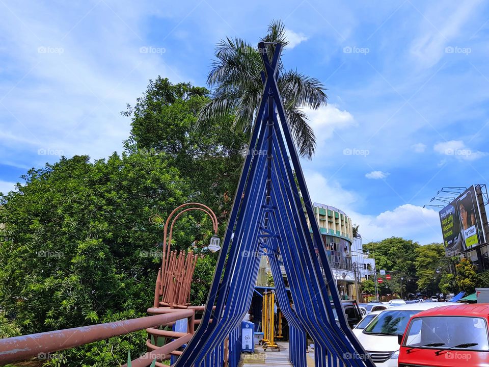 A triangular ornate monument on sidewalk of the sity road, monument is made steel bar, its blue color, vertical below and tilted above to form a triangle, placed in a row, view sky refract blue and clouds are white