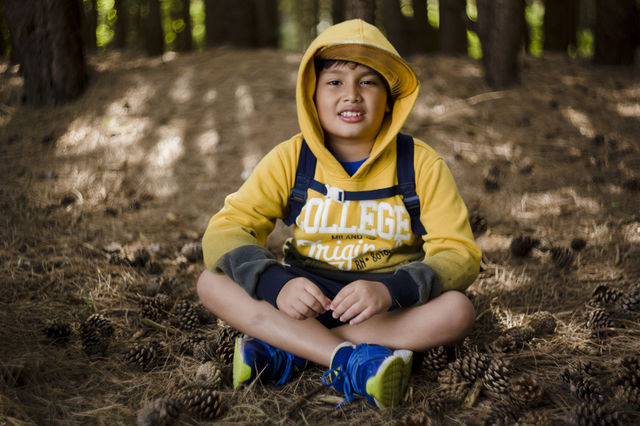 Seasonal outdoor portrait of a young happy Eurasian kid hiking in a pine forest wood.Natural setting, the boy is wearing blue sneakers, yellow sweater, hood and cap, smiling while looking at the camera, seating on the floor surrounded by pinecones