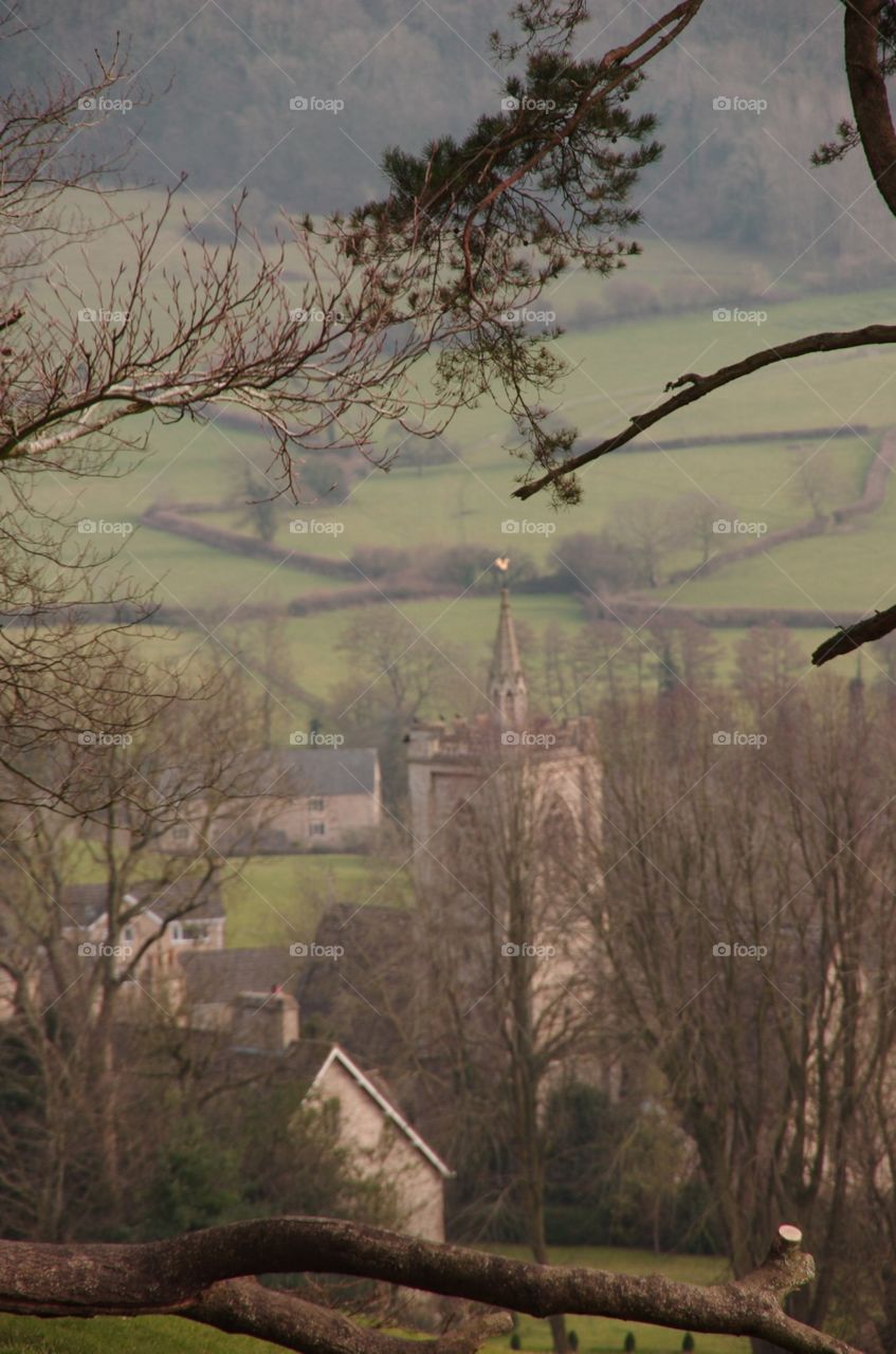 Exterior daylight.  Telephoto shot.  Trees frame the view looking down on Uley’s church.  Background:  bucolic countryside of green fields and stone fences.