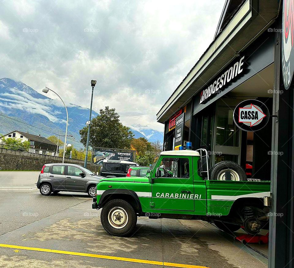 Green color. In the foreground is a green Carabinieri nature conservation car without a rear wheel. There is a wheel in the trunk. In the background are mountains that merge with white clouds.