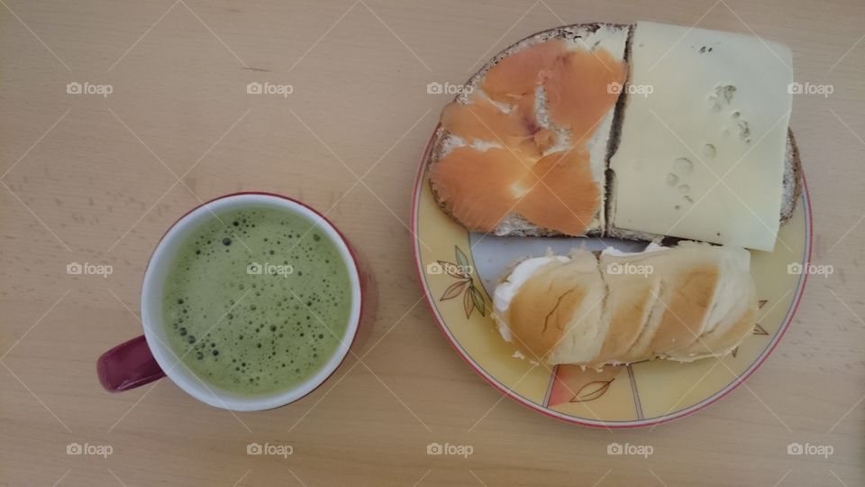 A meal consisting of a cup of matcha green tea, two slices of bread with butter and as topping salmon and cheese respectively, bun with whipped cream as filling on a wooden surface