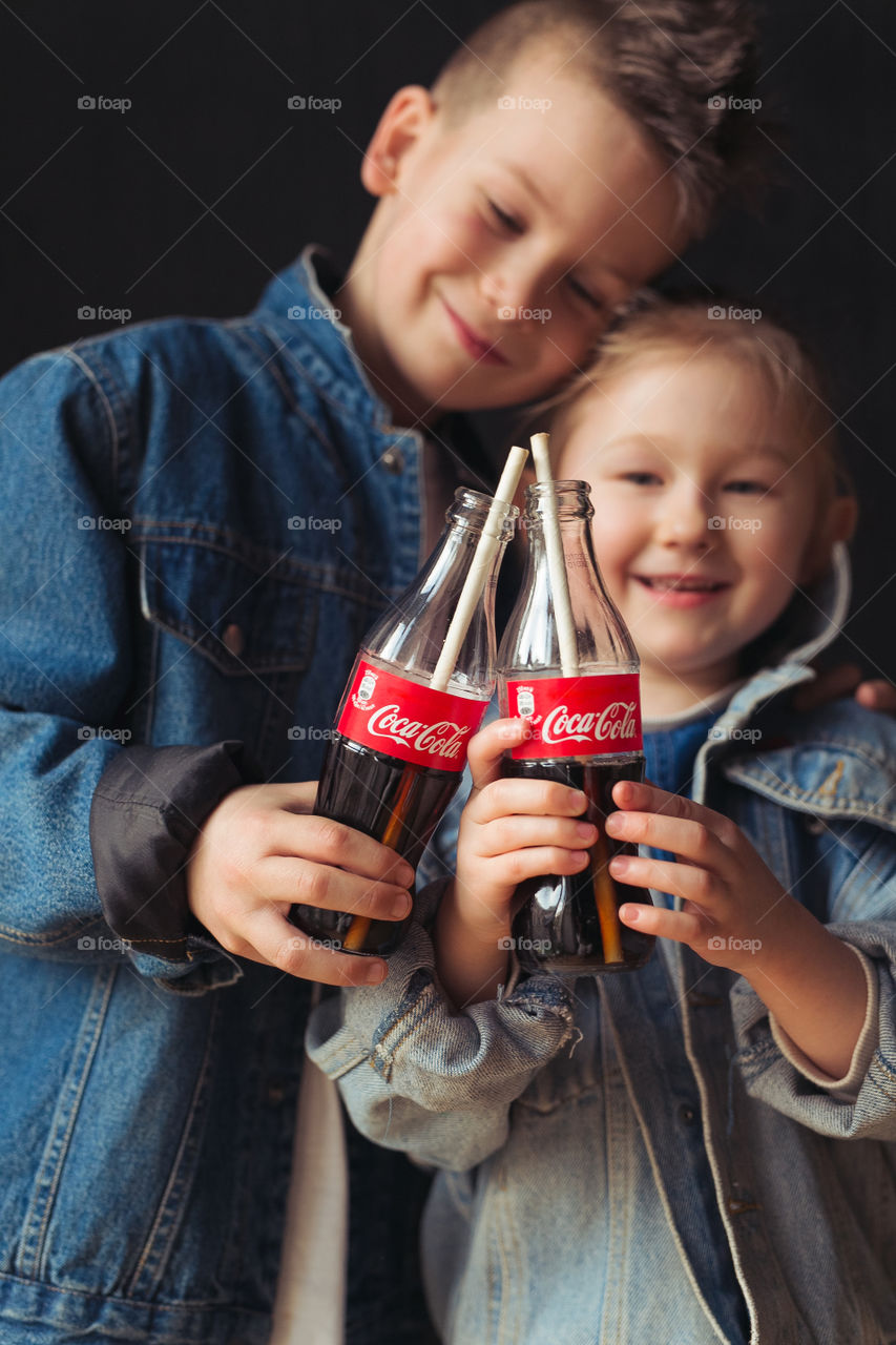 brother and sesira, stay at home, dressed in denim clothes, drink Coca-Cola from bottles through a straw. beautiful stylish kids