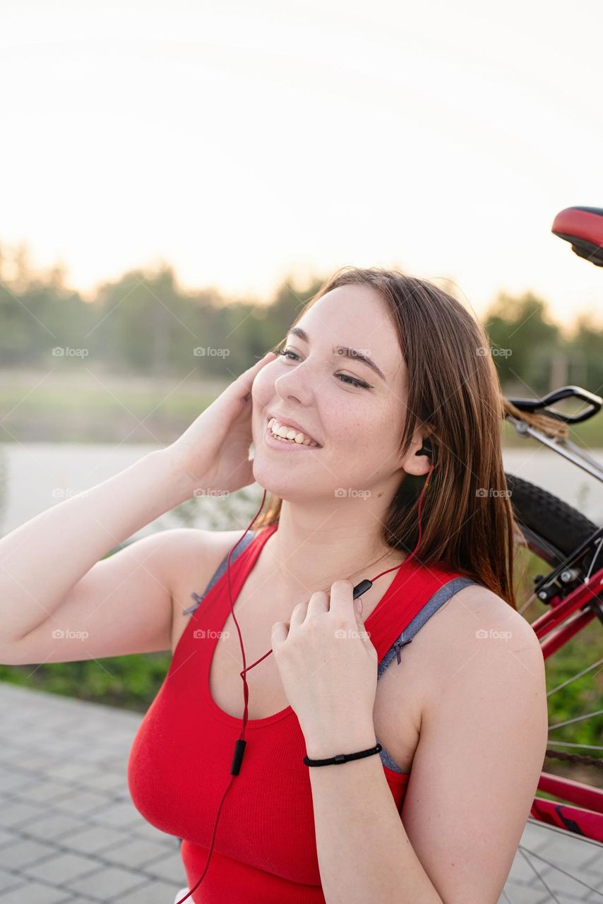 young smiling woman in red shirt and white pants listening to the music standing near her bicycle