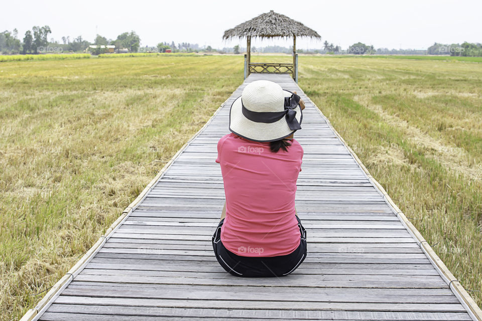 Woman wearing Hat sitting on a wooden bridge with a bamboo hut in the rice fields.