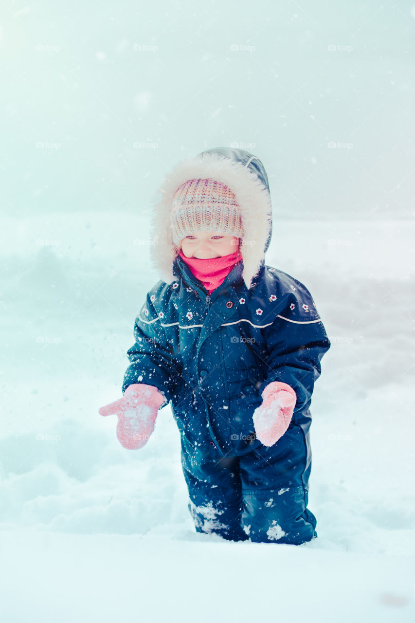 Happy little girl enjoying snow. Child playing outdoors walking through deep snow in wintertime while snow falling. Toddler is wearing dark blue snowsuit