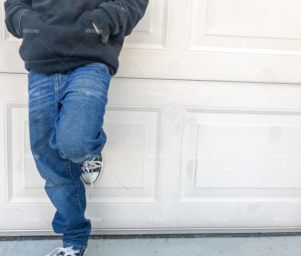 Boy wearing blue jeans and black sweater leaning against wall with one leg bent 