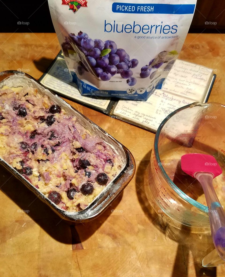 Blueberry Bread preparations, ready to bake in oven. Recipe book, blueberries, mixing bowl & spatula on chopping block.