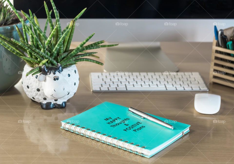 Desk with stationary, computer, pot plant and “Happy Thoughts & Positive Plans” journal